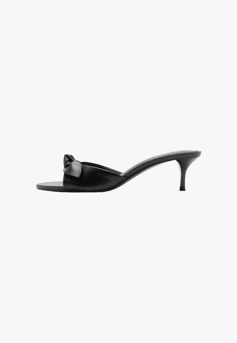 Mules negros de cuero sin cordones con un lazo decorativo, punta abierta y un tacón alto delgado. Textura suave y diseño minimalista.
