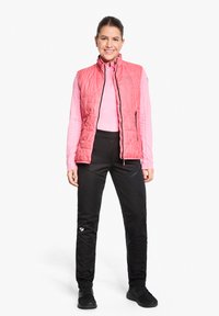 Rosa, gesteppte ärmellose Weste über einem hellrosa Langarmtop, kombiniert mit schwarzen, taillierten Hosen mit kleinem Logo und strukturierten Akzenten.