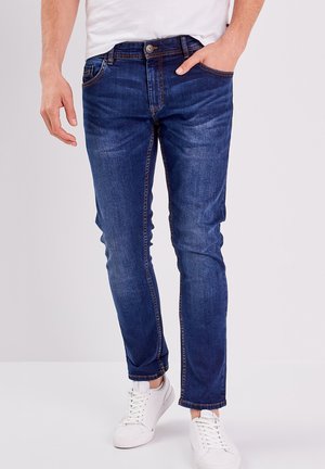 Jeans de mezclilla azul oscuro con un corte ajustado, que presentan detalles de desgaste, cinco bolsillos y costuras en contraste. Combinados con una camiseta blanca y zapatillas.