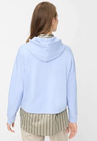 Felpa corta azzurro chiaro con un ampio cappuccio, polsini a costine e una finitura testurizzata, indossata sopra una camicia a righe in tonalità neutre.