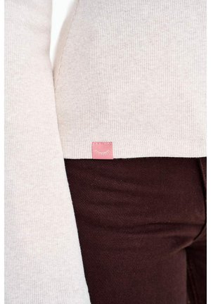 Inimese lähivõte, kes kannab helebeeži soonikuga kampsunit, millel on väike roosa silt, ning tumedat pruunid püksid.