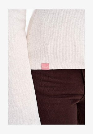 Zbliżenie osoby ubranej w jasnobeżowy prążkowany sweter z małą różową metką i ciemnobrązowe spodnie.