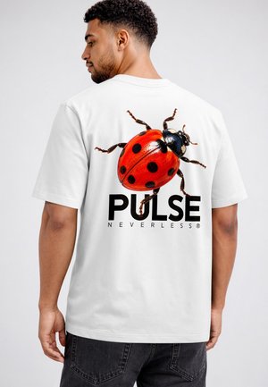 Mann trägt ein weißes T-Shirt mit einer großen rot-schwarzen Marienkäfer-Grafik und den Worten "PULSE NEVERLESS" auf dem Rücken.
