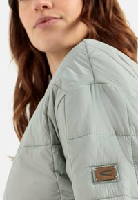 Padded, hellgrüne Jacke aus strukturiertem Stoff mit einem quadratischen Steppmuster und einem braunen Logo-Patch am linken Arm.