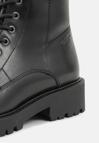 Schwarze Lederstiefel mit einer robusten Sohle, dickem Absatz und Schnürdesign mit Metallösen. Subtile Markenprägung an der Seite.