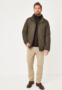 Olivgrüne Jacke mit Reißverschluss, hohem Kragen und Brusttasche, kombiniert mit beigen Hosen und braunen Schuhen. Schlichtes Design, lässige Passform.