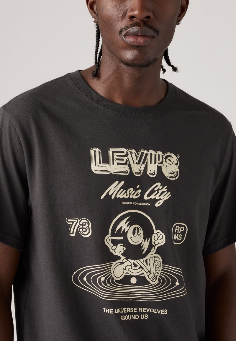 Hombre con camiseta negra Levi's con "Music City", un personaje de vinilo caricaturesco, y el texto "El universo gira a nuestro alrededor".