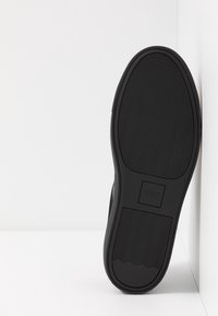 Musta kengänpohja teksturoidusta kumista, jossa on aaltomainen kuviointi ja hillitty logon yksityiskohta keskellä. Kulmikas reuna kohtaa seinän.