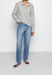 Maglione grigio lavorato a maglia con collo a zip, abbinato a jeans azzurri dal taglio rilassato e sneaker bordeaux con dettagli bianchi.
