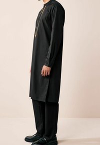 Tunique longue noire à col montant, devant boutonné avec accent doré. En tissu texturé, portée avec un pantalon et des chaussures noirs.