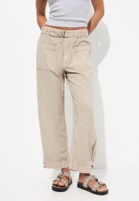 Pantalons beige à jambes larges avec une finition texturée, dotés de deux grandes poches arrière et d'une taille ceinturée, accompagnés de sandales marron à pois.