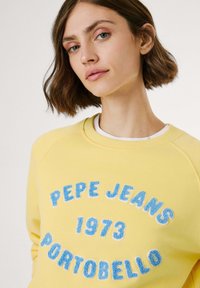 Geltonas džemperis su mėlynu tekstūruotu užrašu "PEPE JEANS 1973 PORTOBELLO." Sukurtas su apykakle ir juostuotais rankogaliais.