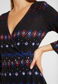 Vestido de punto negro con un profundo escote en V, mangas largas y patrones geométricos en rojo, azul y blanco. Superficie texturizada con un ligero brillo.