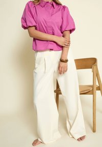 Donna che indossa una blusa rosa acceso con maniche a sbuffo e pantaloni bianchi a gamba larga a vita alta, in piedi a piedi nudi accanto a una sedia di legno.