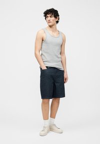Junger Mann steht mit den Händen in den Taschen, trägt ein graues Tanktop, marineblaue Shorts mit Nadelstreifen, weiße Socken und beige Turnschuhe vor einem weißen Hintergrund.