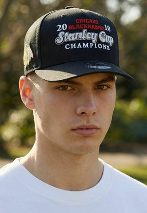 Jeune homme en plein air portant une casquette noire avec une broderie "2010 Chicago Blackhawks Stanley Cup Champions" et un t-shirt blanc à col rond.