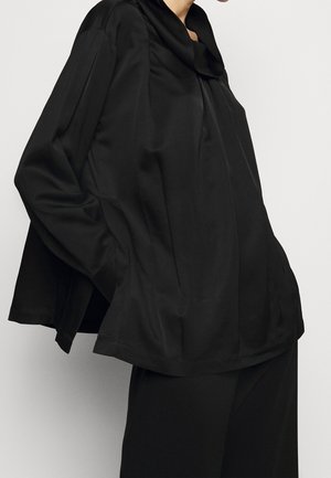 Person trägt eine locker sitzende schwarze Langarmbluse mit weitem Kragen und passende schwarze Hose, steht vor einem schlichten hellen Hintergrund.