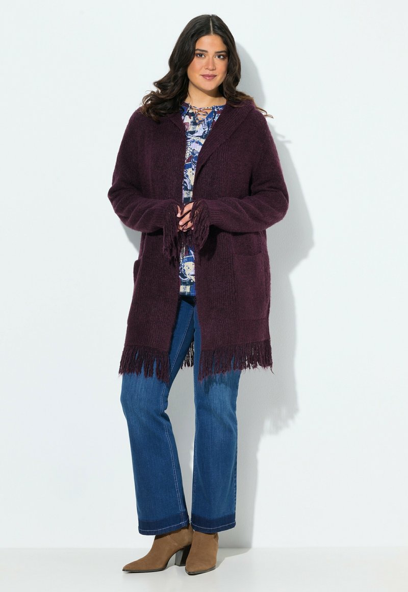 Pflaumenfarbener Strickcardigan mit Fransenumschlag und offener Vorderseite, getragen über einem bedruckten Oberteil und kombiniert mit blauen Jeans und braunen Stiefeletten.