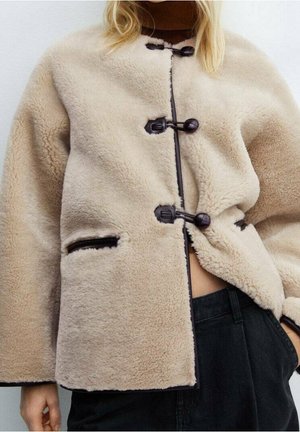 Beige fluffy jakke med toggle-lukninger og lynlåslommer båret over en brun rullekrave, kombineret med sorte bukser.