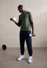 Olivengrønn tettsittende t-skjorte, marineblå joggebukser, hvite treningssko og sølvmanualer. Glatte teksturer i et minimalistisk gymsmiljø.