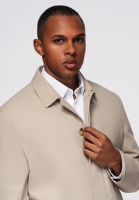 Giacca beige con collo a camicia, chiusura con bottoni e tessuto leggero, indossata sopra una camicia bianca con bottoni.