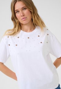 T-shirt en coton blanc avec des embellissements en clous et en œillets argentés sur le devant. Encolure ronde avec des manches courtes, coupe décontractée.