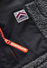 Black jacket with gray fleece lining, Superdry Mountain Sport logo patch, and bright orange Superdry zipper pull tab.