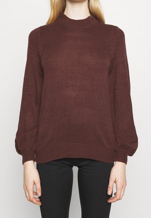 Frau trägt einen langärmeligen, gerippten, dunkelbraunen Strickpullover mit hohem Kragen und Ballonärmeln, kombiniert mit schwarzen Hosen.