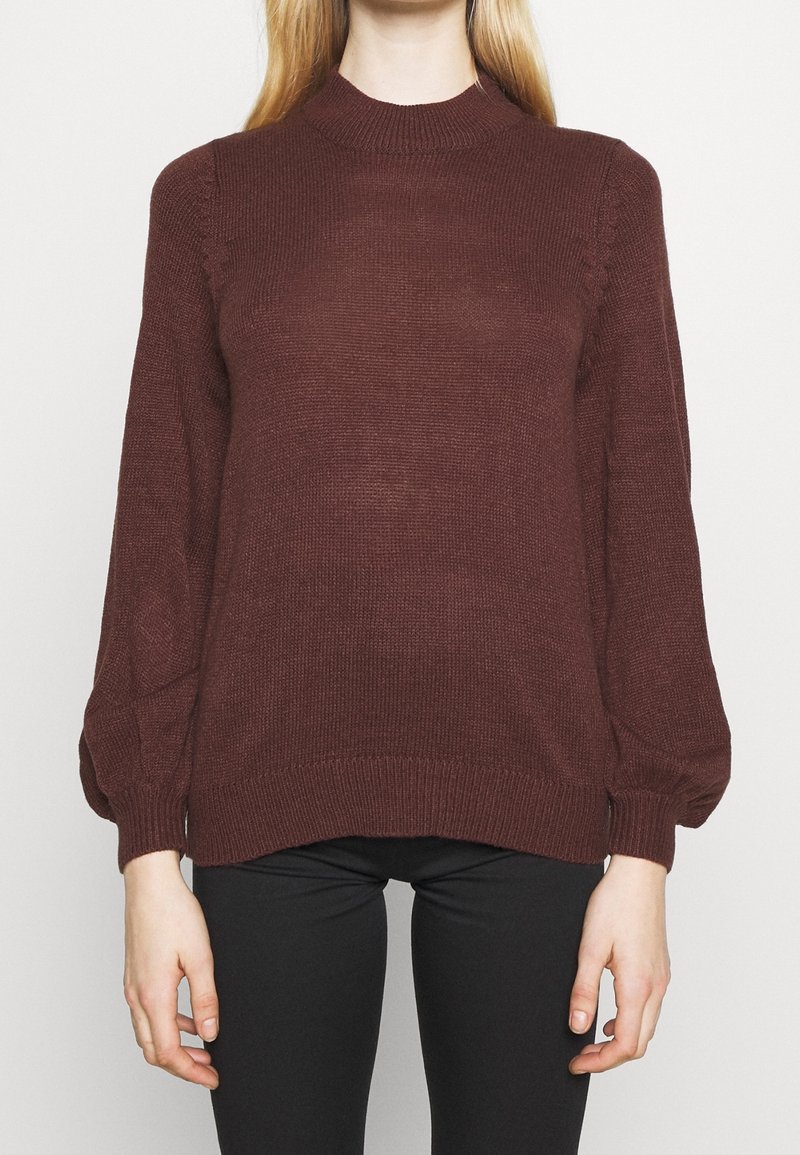 Femme portant un pull en maille côtelé marron foncé à manches longues avec un col montant et des manches ballons, associé à un pantalon noir.