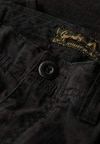 Zwarte stof met een gestructureerd oppervlak, voorzien van een ronde zwarte knop en een groene label met de tekst "Superdry Utility Wear." Close-up detail.