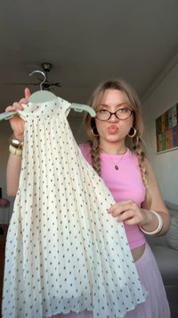 Vestido blanco sin mangas con pequeños lunares negros, diseño plisado y cuello alto, exhibido en un perchero de color azul claro.