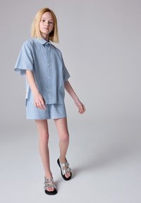 Plava i bijela prugasta košulja kratkih rukava s odgovarajućim kratkim hlačama, uparena s metalik sandalama s podesivim kopčama i teksturiranim đonom.