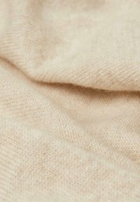Tissu en tricot beige, texture douce, montrant de fines fibres et un motif de tissage légèrement irrégulier. Le tissu se drape doucement avec une élasticité visible.