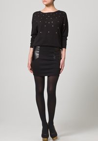 Schwarzer Strickpullover mit goldenen Nieten, kombiniert mit einem schwarzen Minirock mit Kunstlederpaneelen. Das Modell trägt schwarze Strumpfhosen und Schuhe.