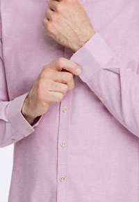 Camisa de manga larga rosa claro con tejido texturizado, cierre de botones en la parte delantera y puños con un solo botón. Presenta un cuello clásico y botones redondos.