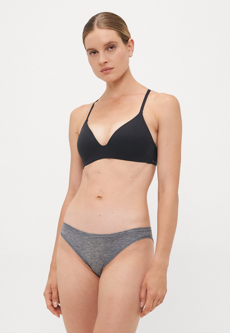 Woman with pulled-back hair wearing a black bra and grey underwear, standing against a plain light background.