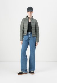 Giacca verde oliva imbottita con colletto alto, camicia nera con bottoni, jeans blu in denim a gamba ampia e sneakers nere con suola in gomma.