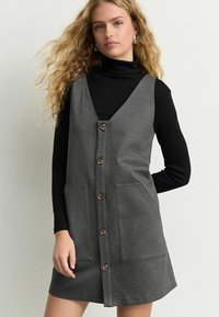 Halli sansleeve kleit, nööpidega ees, kahe ees oleva taskuga. Kanda musta kõrge kaelusega pluusi peal, millel on sile tekstuur ja minimalistlik disain.