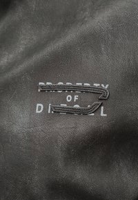 Μαύρη υφή δέρματος με μερικώς ορατό λευκό κείμενο "PROPERTY OF DIESEL" και δύο επικαλυπτόμενα ραμμένα δερμάτινα μπαλώματα.