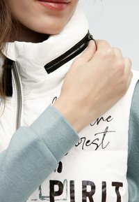 Weißes gepolstertes Vest mit hohem Kragen, ausgestattet mit einem schwarzen Reißverschluss und grafischen Textdetails. Ein hellblaues Langarmhemd ist teilweise darunter sichtbar.