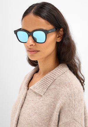 Donna con lunghi capelli scuri, che indossa occhiali da sole blu specchiati e un maglione beige lavorato a maglia con un ampio colletto, guarda leggermente di lato.