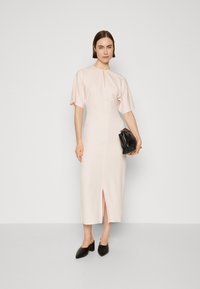 Vaaleanpunainen istuva mekko lyhyillä hihoilla, jossa on etuhalkio ja avonainen kaula-aukko. Yhdistetty mustaan clutch-laukkuun ja korkokenkiin.