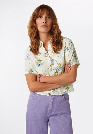 Kortærmet, skjorte med knapper i lyst beige stof med et farverigt blomstret og tropisk print; kombineret med højtaljede lavendel-jeans.