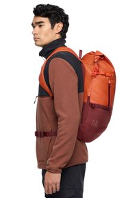 Mochila naranja y roja con un acabado texturizado, correas ajustables y un bolsillo lateral, llevada sobre una chaqueta de forro polar marrón.