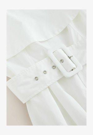 Curea din material textil alb trecută printr-o cataramă dreptunghiulară cu inele metalice, pe un articol vestimentar alb.