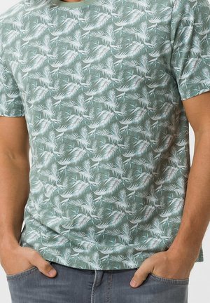 Homme portant un t-shirt vert à motif de feuilles de palmier tropicales blanches, les mains dans les poches, associé à un jean bleu.