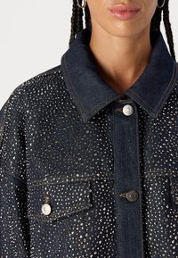 Chaqueta de mezclilla con un acabado oscuro, adornada con rhinestones plateados, con cuello tipo spread, bolsillos frontales y cierres de botón metálico.