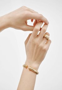 Joyería de metal en tono dorado que incluye un anillo en espiral y una pulsera de puño a juego. Textura suave con acentos estriados. Manos que ajustan el anillo.