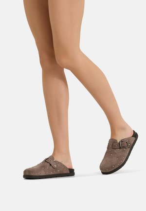 Gambe che indossano sabot scamosciati taupe slip-on con piccole borchie, cinturino regolabile con fibbia e suole nere, mostrati su sfondo bianco.