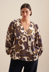 Una blusa leggera presenta un vivace motivo paisley in blu navy e oro su uno sfondo bianco. La scollatura a V e le maniche a sbuffo valorizzano il design.